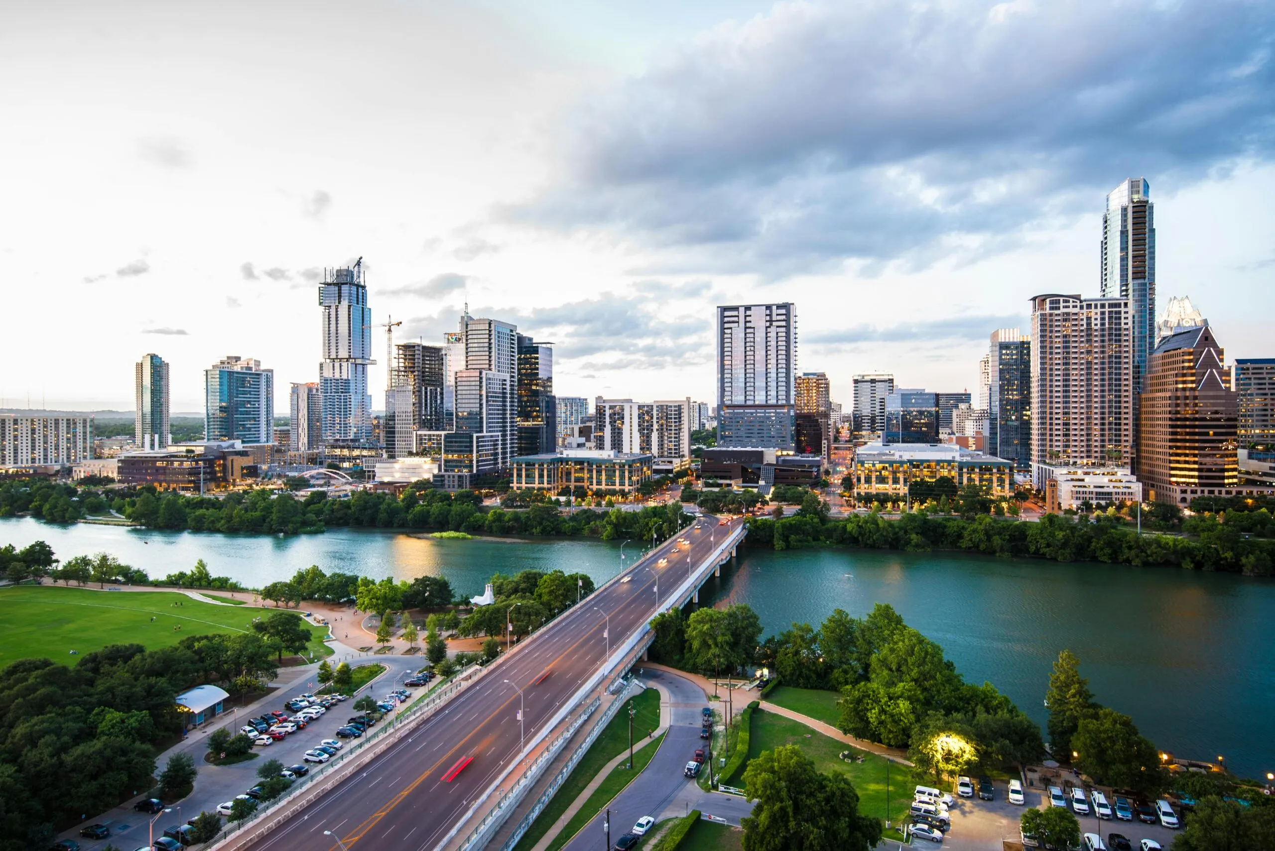 Insights Blog 1 Aerial view of downtown Austin, Texas, featuring modern skyscrapers, the Colorado River, a bridge connecting the city center, and lush green parkland under a cloudy evening sky.