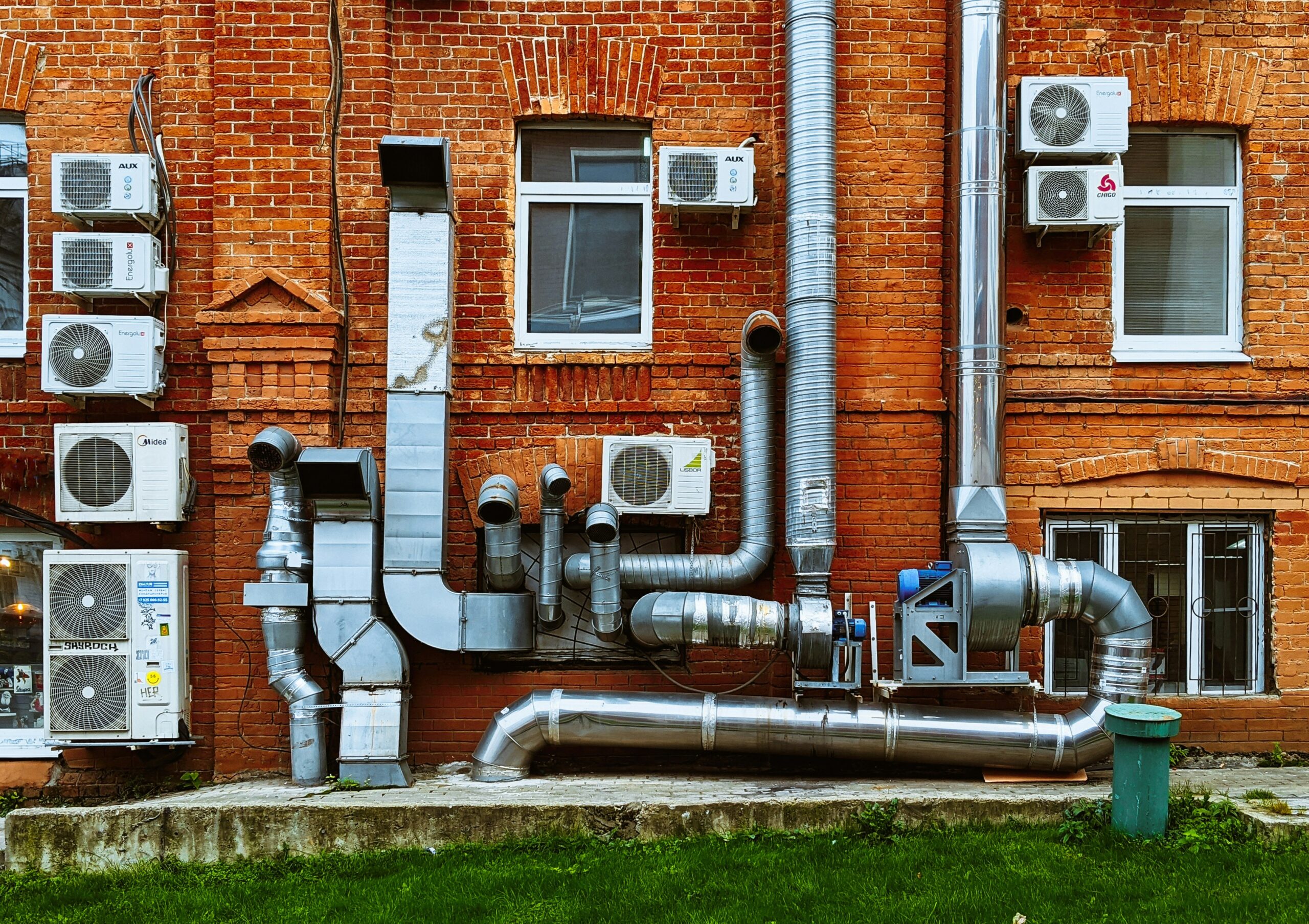 Exterior wall of a commercial brick building fitted with multiple HVAC condenser units, large metal ventilation ducts, exhaust pipes, and industrial air-handling equipment used for climate control and air circulation.
