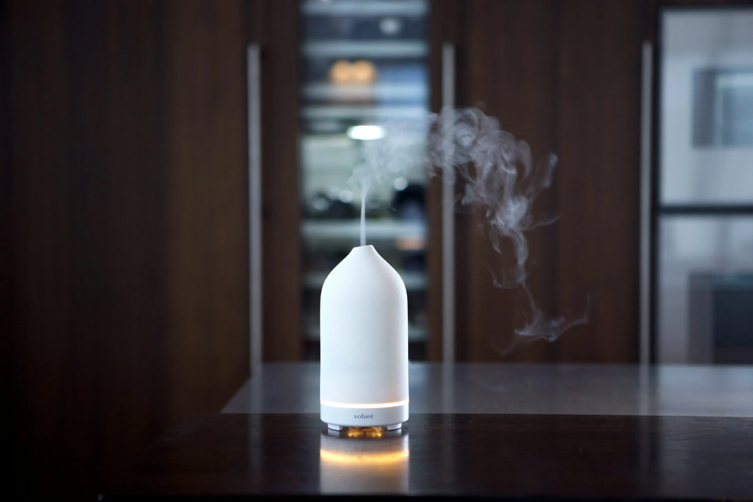 Insights Blog 7 A compact white ultrasonic humidifier releasing a stream of cool mist on a dark tabletop in a modern indoor setting, demonstrating how a humidifier adds moisture to dry indoor air.