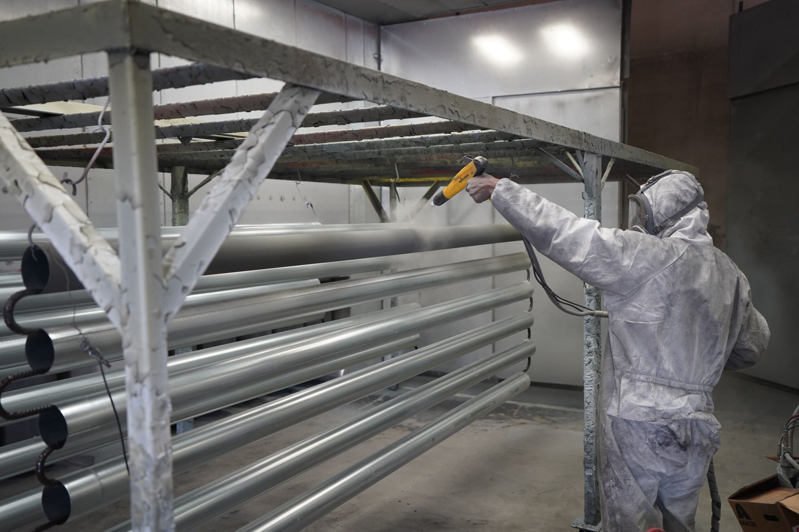 Insights Blog 9 Industrial worker applying a protective powder coating to metal tubing in a manufacturing facility using a spray gun and protective equipment.