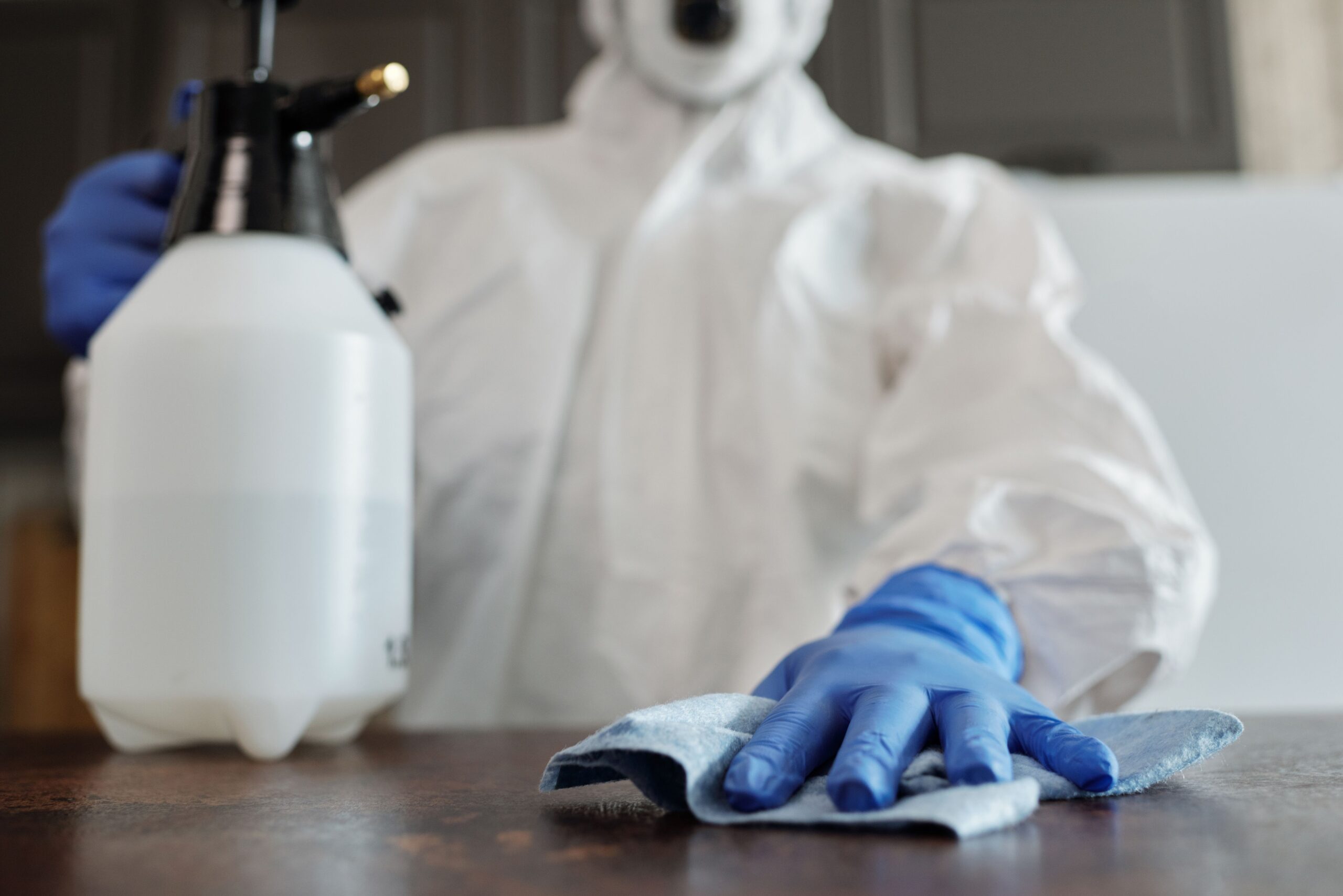 Insights Blog 6 Person wearing protective coveralls, gloves, and a face mask disinfects a tabletop surface with a cleaning cloth while a spray bottle of disinfectant sits nearby.