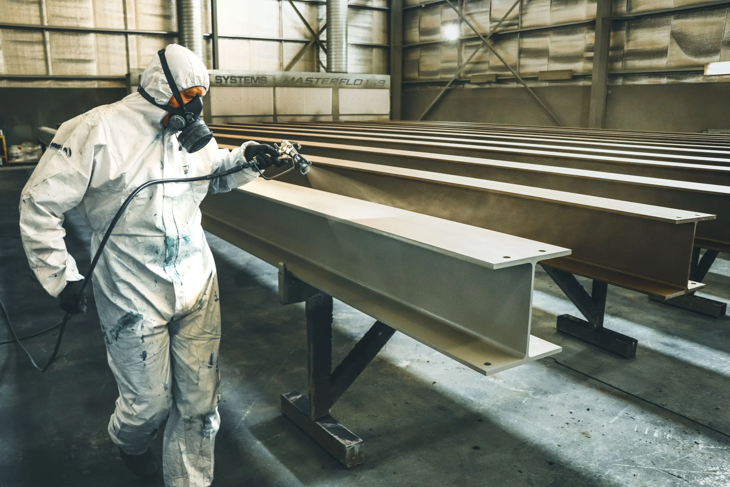 Insights Blog 2 Worker in protective coveralls and respirator spray-coating large steel beams inside an industrial paint booth, illustrating paint application processes where static control helps reduce overspray, dust contamination, and finish defects.