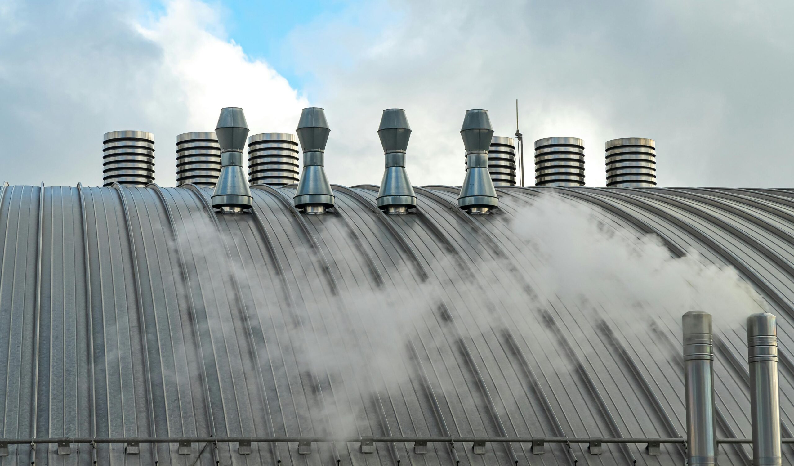 Insights Blog 9 Industrial building rooftop with steam exhaust and ventilation stacks releasing water vapor, illustrating large-scale environmental control systems used in manufacturing facilities and commercial HVAC operations.