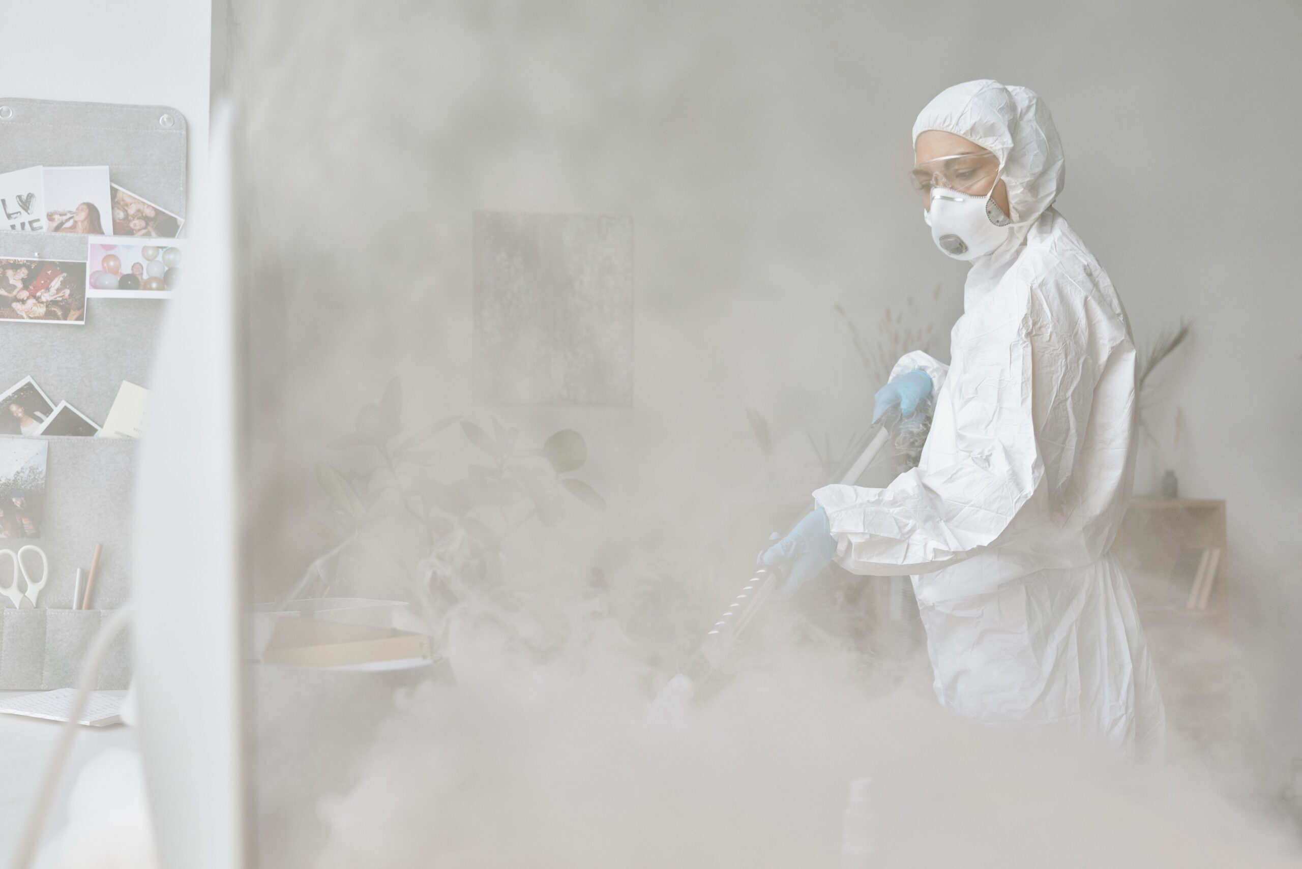 Insights Blog 3 Worker wearing protective coveralls, gloves, goggles, and a respirator operating a fogging machine in an indoor commercial space, illustrating chemical disinfection fogging used for facility sanitation and pathogen control.