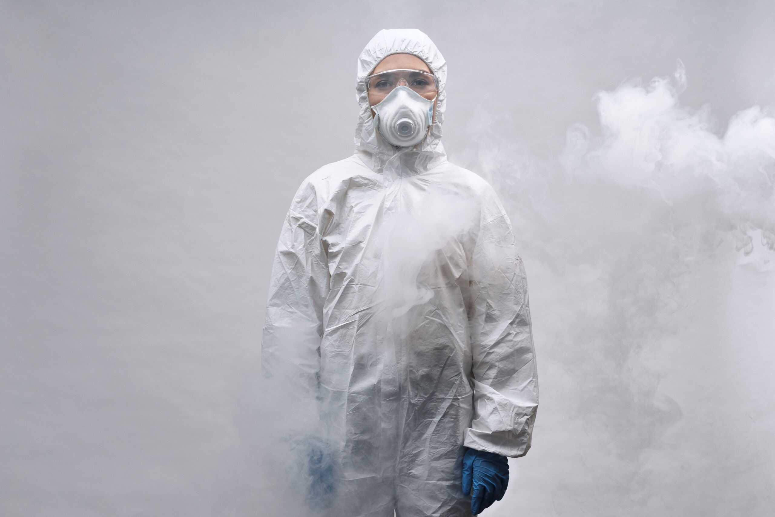 Worker wearing protective coveralls, safety goggles, and a respirator standing in a fog-filled industrial environment to illustrate airborne dust and contaminant exposure.