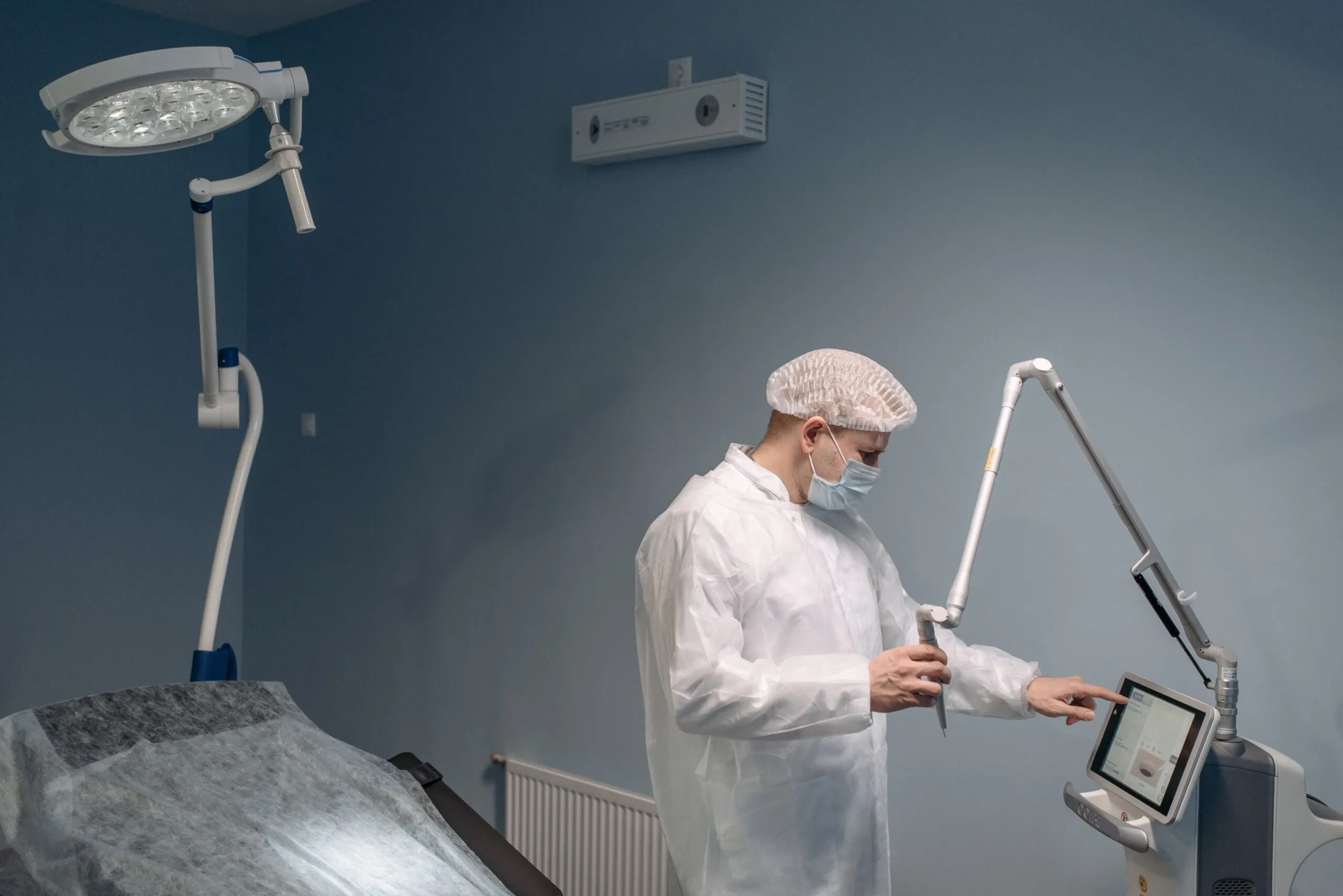 Gowned technician adjusts a wall-mounted control panel monitoring operating room humidity, with surgical lights and ceiling air unit nearby.