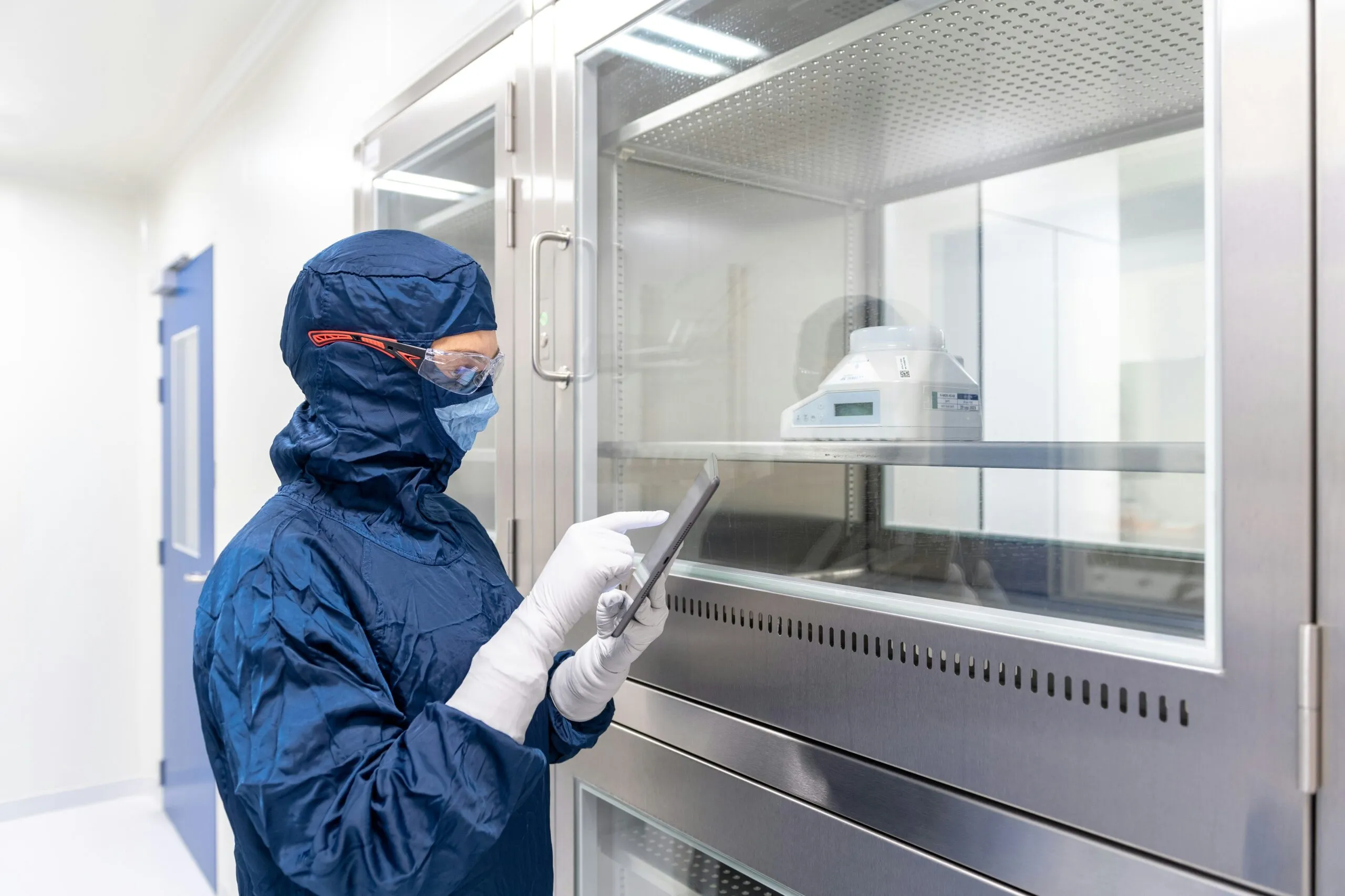 A technician in full cleanroom gowning, including a hood, safety glasses, and mask, checks a tablet device in front of a sealed equipment enclosure inside a controlled pharmaceutical or cleanroom facility.